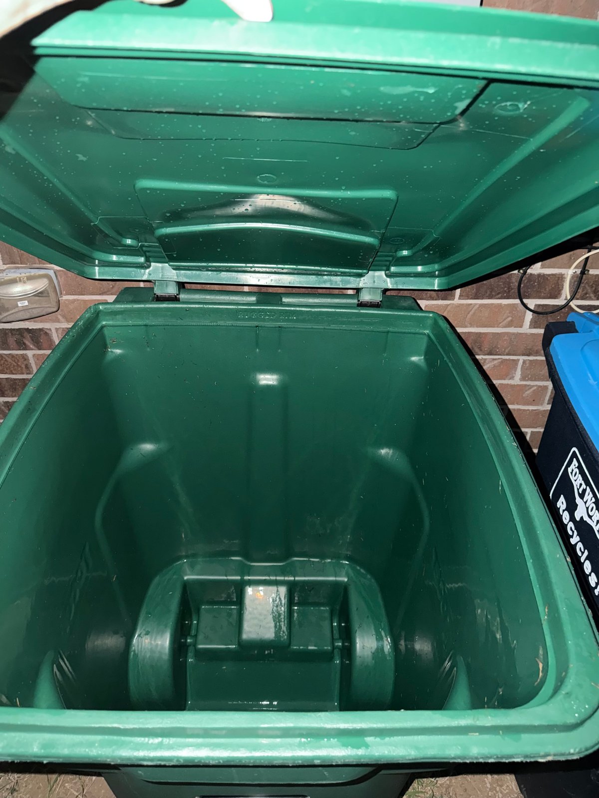 Open green waste bin with lid raised, showing empty interior and wheel at bottom, against brick wall