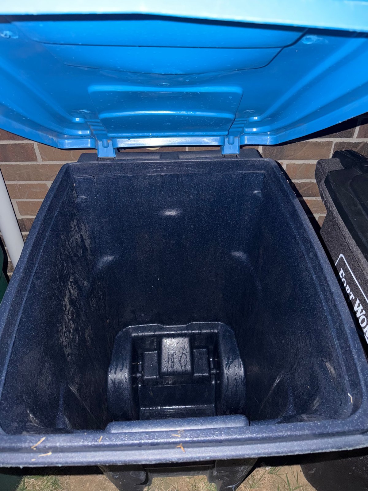 Open blue and black trash bin showing empty dark interior with locking mechanism at bottom and brick wall background